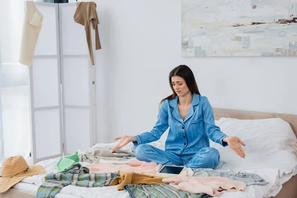 Mulher descontente no elegante pijama apontando para muitas roupas na cama perto de tablet digital com tela em branco — Fotografia de Stock