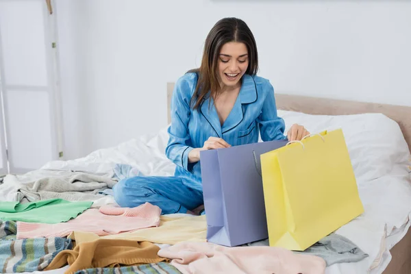 Fröhliche Frau blickt in Einkaufstüten neben viel Kleidung im Schlafzimmer — Stockfoto