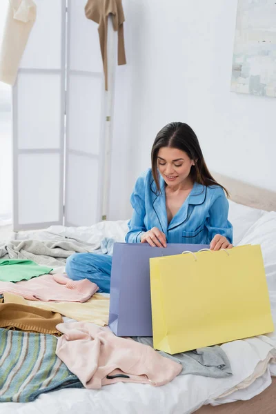 Femme heureuse en pyjama de soie regardant dans les sacs à provisions près de beaucoup de vêtements dans la chambre — Photo de stock