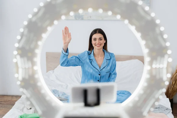 Felice fashion blogger in elegante pigiama agitando mano vicino porta telefono sfocato con lampada ad anello — Foto stock