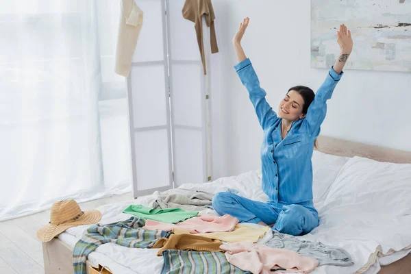 Mujer alegre en pijama de seda estirándose en la cama cerca de un montón de ropa diferente - foto de stock