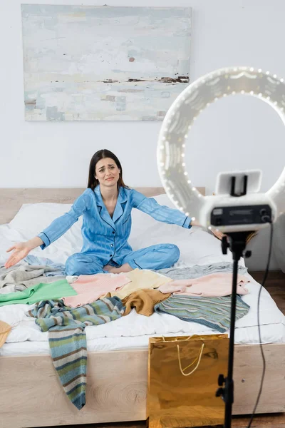 Displeased woman in trendy pajama pointing at plenty of clothes near phone holder with circle light — Stock Photo