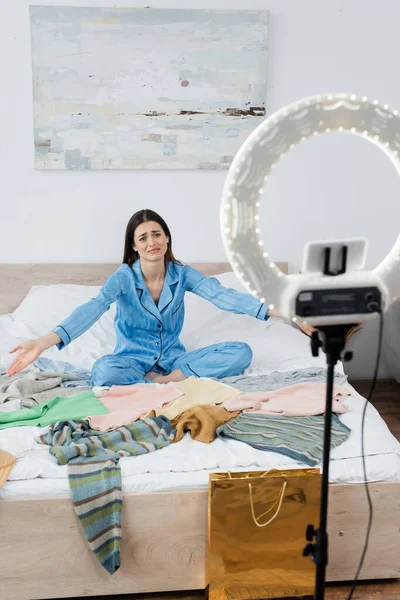 Mulher descontente na moda pijama apontando para uma abundância de roupas perto titular do telefone com luz círculo — Fotografia de Stock