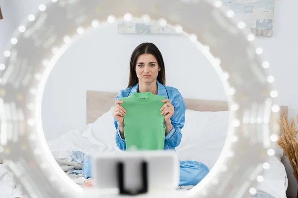 Titular de teléfono borroso con círculo de luz cerca descontento blogger moda mostrando chaleco - foto de stock