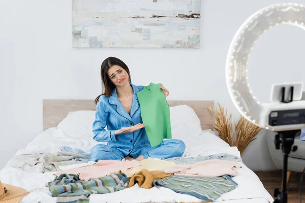 Displeased fashion blogger pointing at vest near clothes on bed and phone holder with ring light — Stock Photo