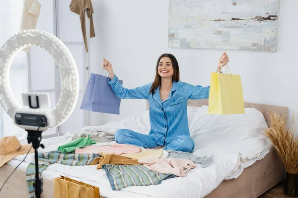 Bloguero de moda feliz con bolsas de compras cerca de surtido de ropa y titular del teléfono con lámpara de anillo - foto de stock