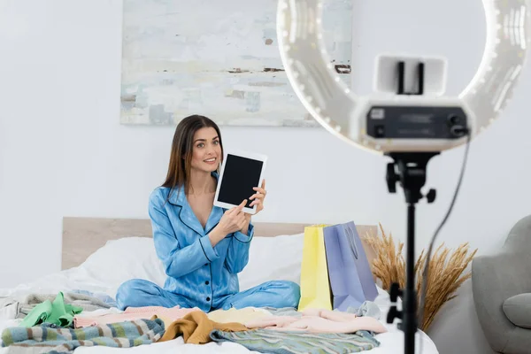 Mujer feliz en pijama de seda apuntando a la tableta digital con pantalla en blanco cerca de la ropa y el titular del teléfono borroso - foto de stock