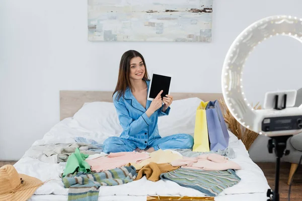 Mulher sorridente em pijama de seda apontando para tablet digital perto de roupas e suporte de telefone borrado — Fotografia de Stock