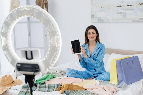 Happy fashion blogger in trendy pajama showing digital tablet near phone holder with ring lamp — Stock Photo