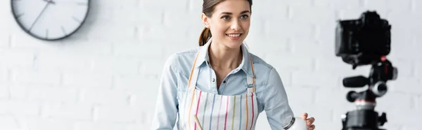 Sonriente blogger culinario sosteniendo salero cerca borrosa cámara digital en la cocina, pancarta - foto de stock