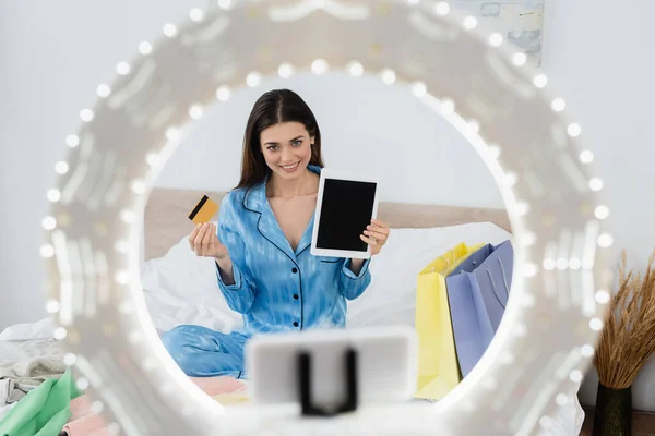 Lächelnder Modeblogger zeigt digitales Tablet und Kreditkarte in der Nähe einer verschwommenen Handyhalterung mit Ringlampe — Stockfoto