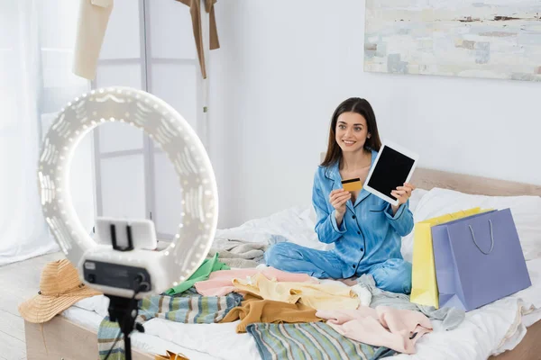 Mujer sonriente en elegante pijama con tableta digital y tarjeta de crédito cerca de la ropa y el titular del teléfono con lámpara de anillo - foto de stock