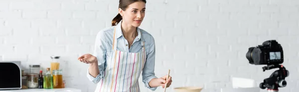 Молодой кулинарный блоггер указывая рукой возле цифровой камеры на кухне, баннер — стоковое фото