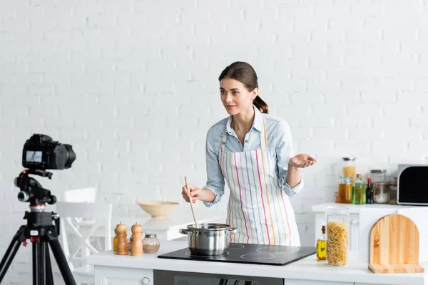 Junge kulinarische Bloggerin spricht in der Nähe der Digitalkamera, während sie in der Küche Essen zubereitet — Stockfoto