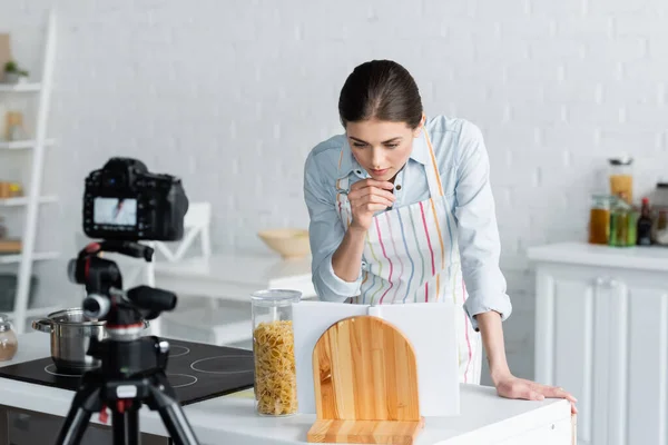 Young housewife looking at cookbook near pasta and blurred digital camera — Stock Photo