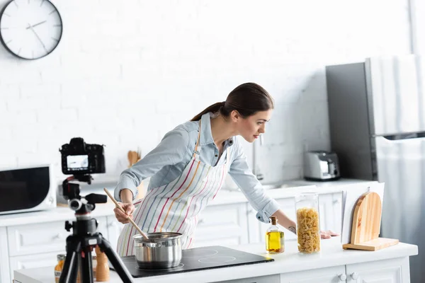 Mujer joven mirando el libro de cocina durante la preparación de alimentos frente a la cámara digital borrosa - foto de stock