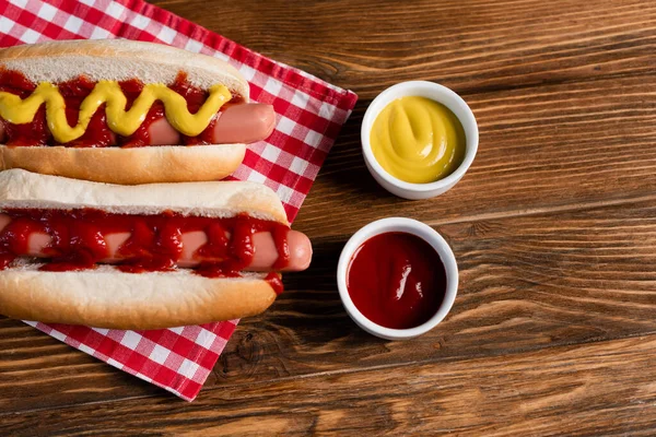 Top view of hot dogs near sauce bowls and checkered napkin on wooden table — Stock Photo