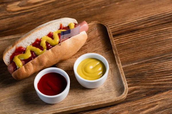 Hot dog con piccola bandiera USA vicino a ciotole con salse sul tavolo di legno — Foto stock