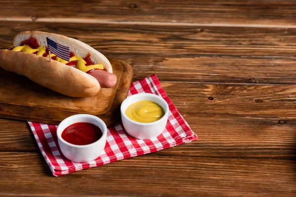 Hot Dog mit kleiner amerikanischer Flagge auf Schneidebrett und Soßen auf karierter Serviette und Holztisch — Stockfoto