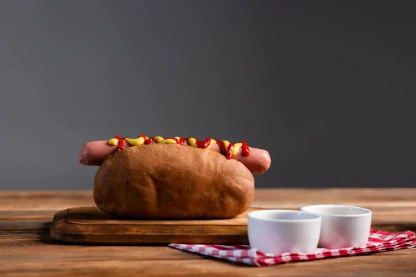 Tazones de salsa y servilleta a cuadros cerca de sabrosos perros calientes en mesa de madera aislada en gris - foto de stock