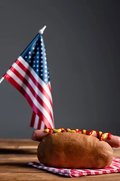 Blurred american flag, hot dog and plaid napkin on wooden table isolated on grey — Stock Photo