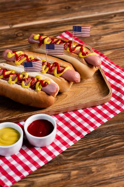 Hotdogs mit kleinen US-Flaggen auf Tablett und karierter Serviette neben Ketchup und Senf auf Holztisch — Stockfoto