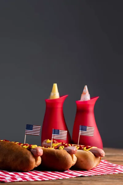 Botellas de salsa borrosa y perros calientes con banderas americanas pequeñas en servilleta de mesa a cuadros aislada en gris - foto de stock