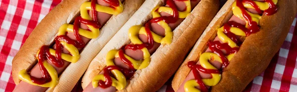 Vista de perto de saborosos cachorros-quentes com mostarda e ketchup na toalha de mesa xadrez, banner — Fotografia de Stock