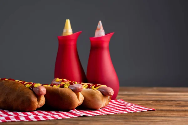 Bottiglie di salsa sfocata vicino gustosi hot dog su tovagliolo plaid isolato su grigio — Foto stock