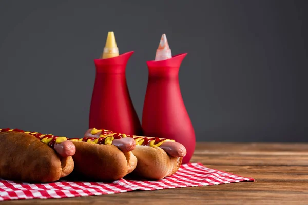 Botellas de salsa borrosa cerca de sabrosos perros calientes en servilleta a cuadros aislado en gris - foto de stock