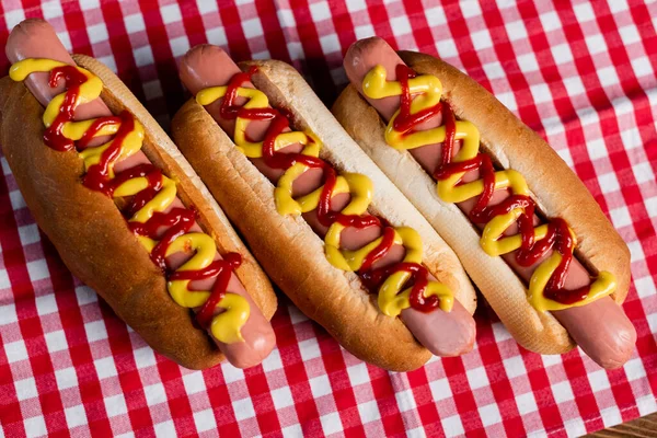 Vista superior de deliciosos cachorros-quentes com mostarda e ketchup na toalha de mesa xadrez — Fotografia de Stock