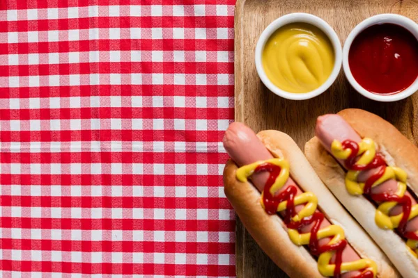 Top view of wooden tray with hot dogs and bowls with mustard and ketchup on plaid tablecloth — Stock Photo