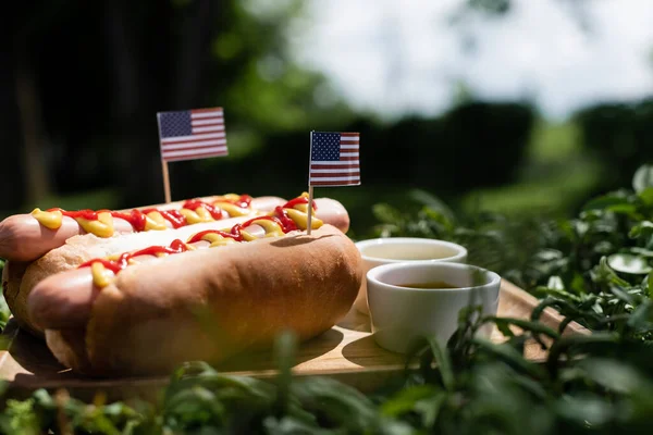 Cachorros-quentes com pequenas bandeiras americanas perto de ketchup e mostarda no gramado verde — Fotografia de Stock