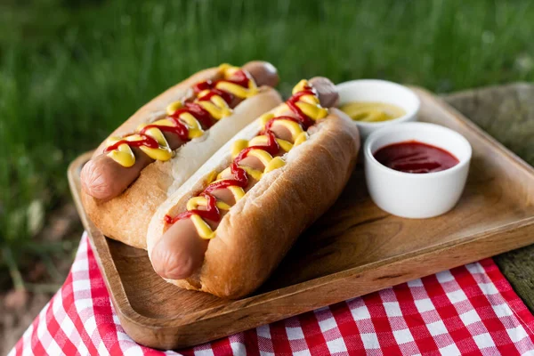 Wooden tray with sauce bowls and testy hot dogs on checkered table napkin outdoors — Stock Photo