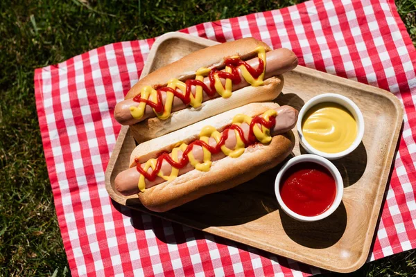 Vista superior de guardanapo xadrez e bandeja de madeira com cachorros-quentes e tigelas de molho no gramado verde — Fotografia de Stock