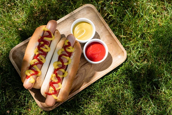 Vista superior de bandeja de madera con hot dogs, mostaza y ketchup sobre hierba verde - foto de stock