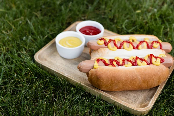 Plateau en bois avec hot dogs, moutarde et ketchup sur pelouse verte à l'extérieur — Photo de stock