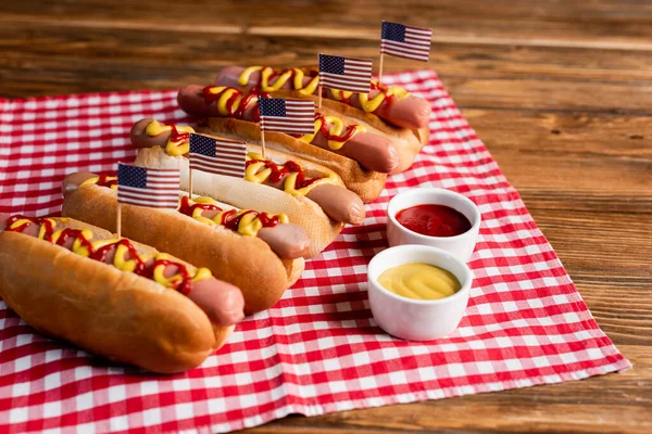 Sabrosos perros calientes con banderas americanas pequeñas cerca de salsas y servilleta a cuadros en mesa de madera - foto de stock