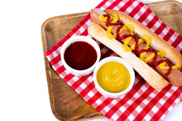 Saboroso cachorro-quente com mostarda e ketchup em guardanapo de mesa xadrez e bandeja de madeira isolada em branco — Fotografia de Stock