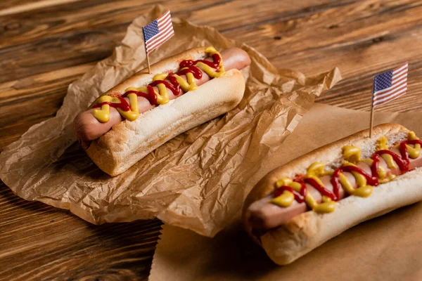 Cachorros-quentes com ketchup, mostarda e pequenas bandeiras americanas em papel kraft e mesa de madeira — Fotografia de Stock