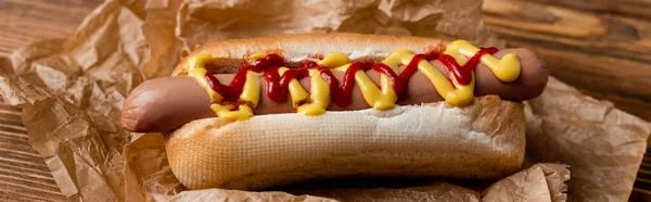 Close up view of hot dog with ketchup and mustard, and crumpled kraft paper on wooden table, banner — Stock Photo
