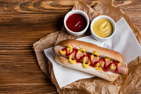 Top view of hot dog near sauce bowls, paper napkin and crumpled kraft parchment on wooden table — Stock Photo