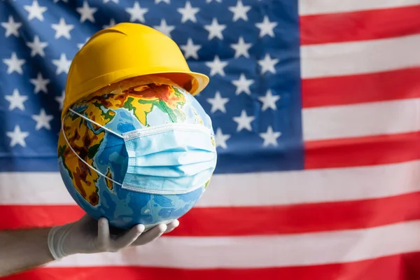 Обрізаний вид людини в латексній рукавичці, що тримає глобус з хардхатом і медичною маскою біля розмитого прапора США, концепція робочого дня — стокове фото