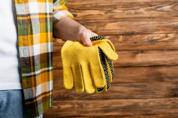 Vista recortada del manitas en camisa a cuadros sosteniendo guantes de trabajo sobre fondo de madera, concepto del día del trabajo - foto de stock