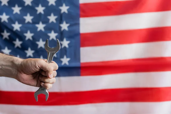 Teilansicht eines Mannes mit Schraubenschlüssel in der Nähe der US-Flagge auf verschwommenem Hintergrund, Konzept für den Tag der Arbeit — Stockfoto