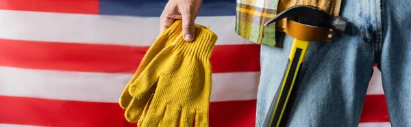 Vista recortada del trabajador con martillo en la correa de herramientas que sostiene los guantes de trabajo cerca de la bandera de EE.UU., concepto del día del trabajo, bandera - foto de stock