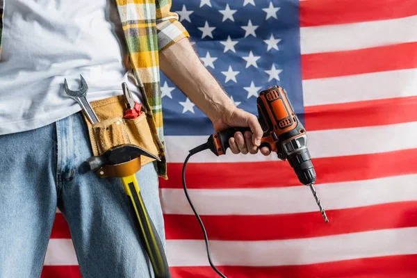 Vue partielle du constructeur dans la ceinture d'outils tenant perceuse électrique près du drapeau des États-Unis, concept de la journée de travail — Photo de stock