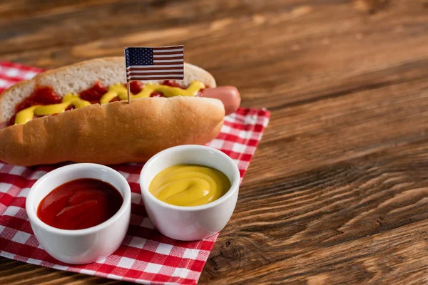 Миски с соусами рядом с вкусным хот-догом с маленьким американским флагом и клетчатой салфеткой на деревянном столе — стоковое фото