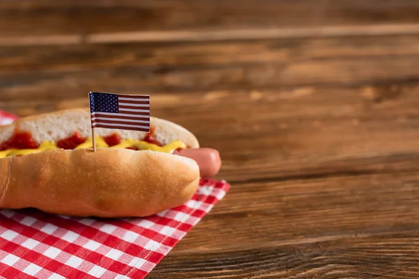 Leckerer Hot Dog mit kleiner amerikanischer Flagge auf karierter Serviette und Holztisch — Stockfoto