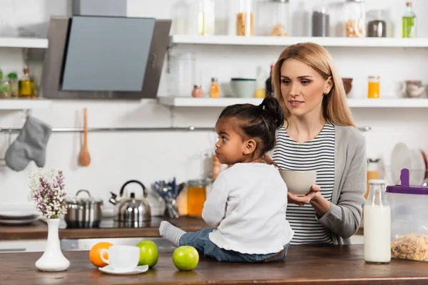 Mulher segurando tigela e olhando adotado garoto americano africano sentado na mesa da cozinha — Fotografia de Stock